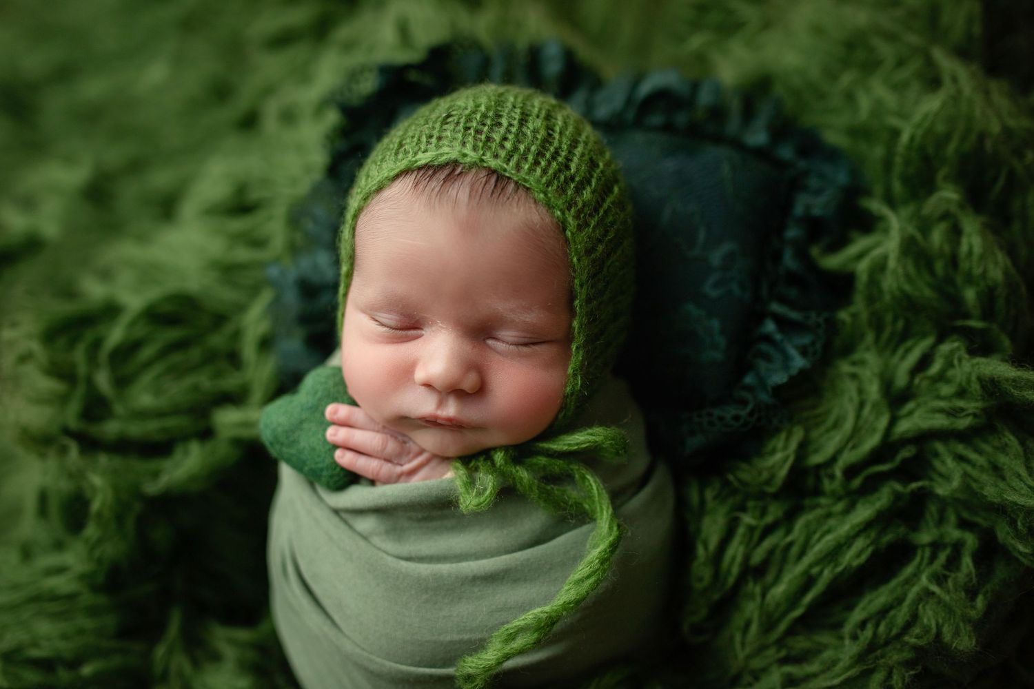 Sleeping newborn in green wrap and knit hat nestled in dark green textured backdrop.