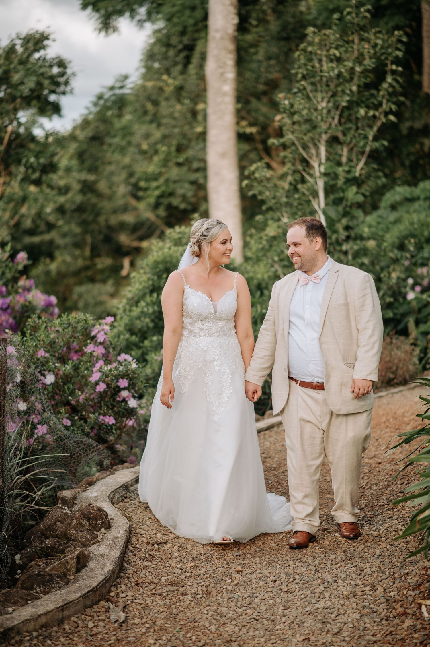 A couple walks hand in hand along a garden path surrounded by blooming flowers and palm trees.