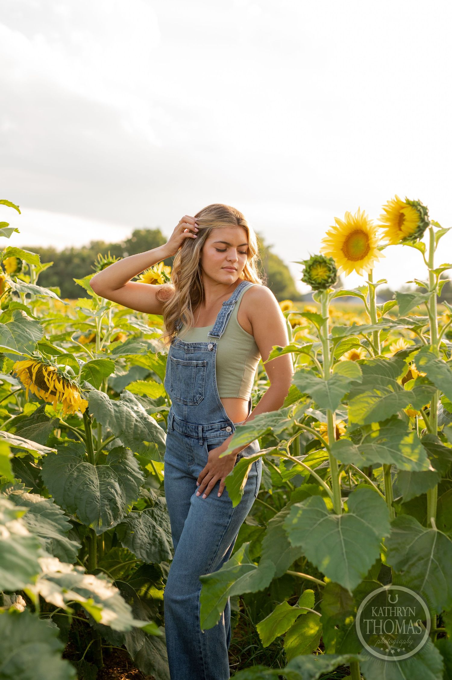 Someone in denim overalls poses casually in a vibrant sunflower field at sunset.