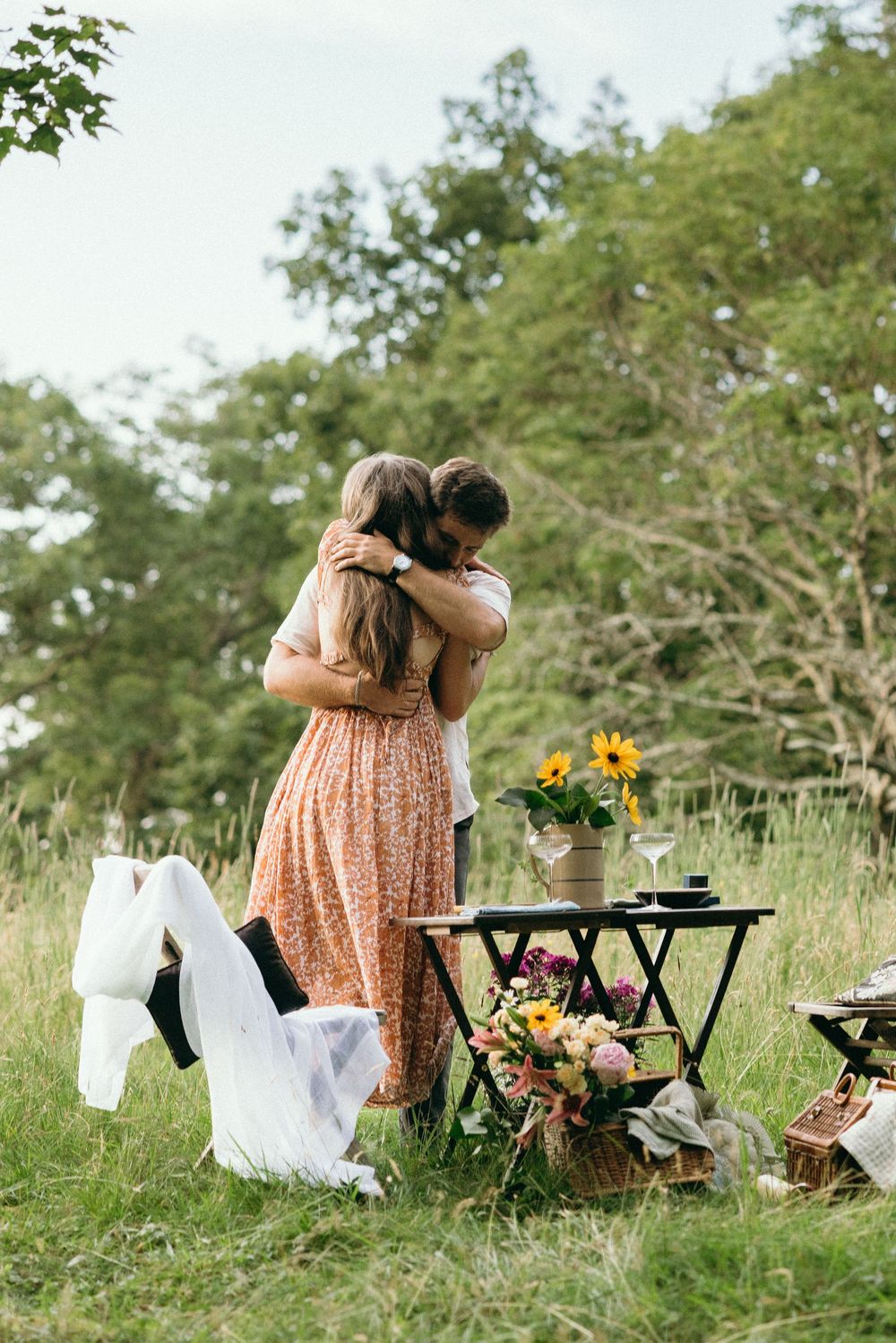 Couple embracing in meadow with picnic table decorated with sunflowers and linens. Luxury Estate Wedding Photographer in NC