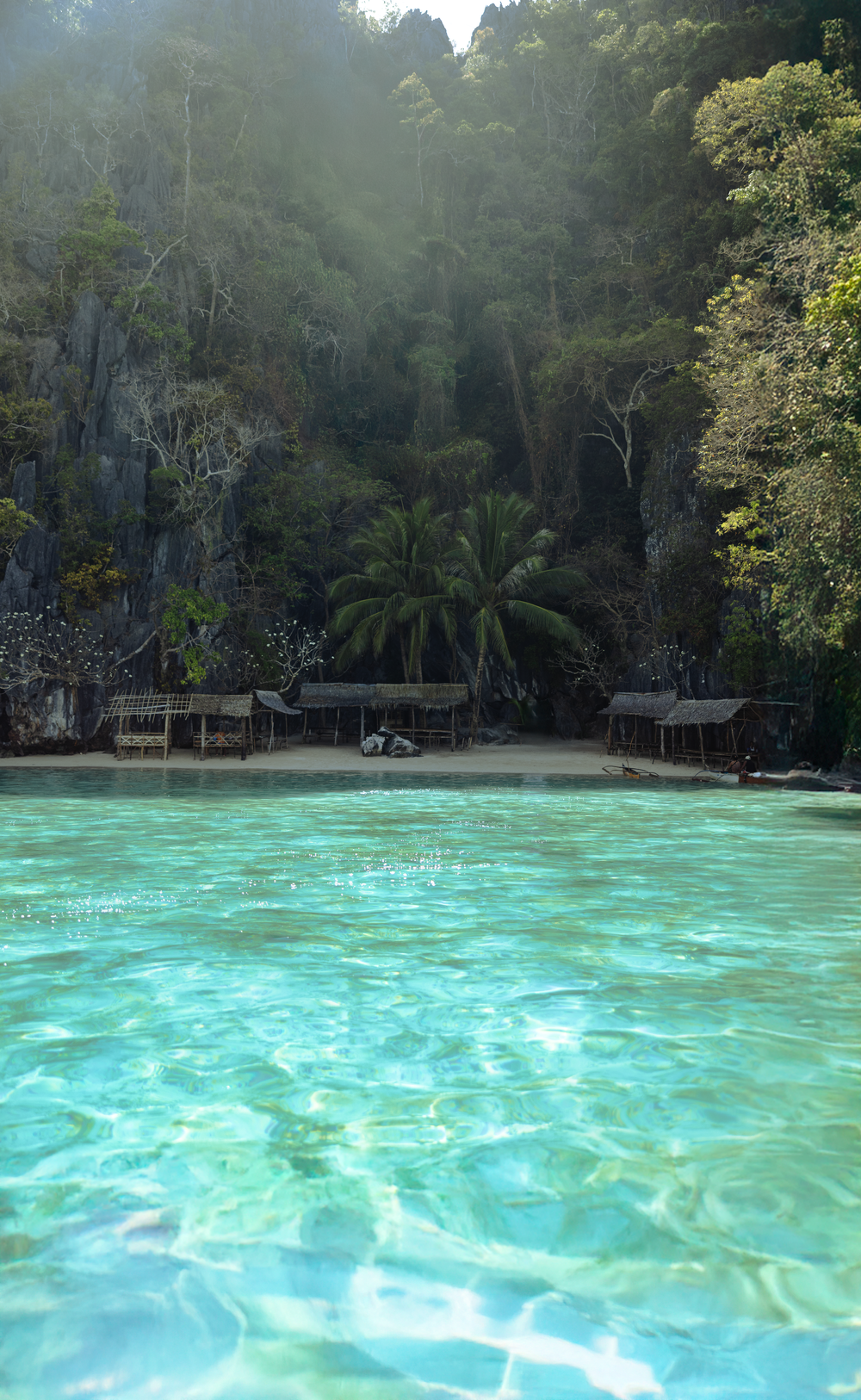 Turquoise waters lap at a secluded tropical beach with wooden huts and limestone cliffs surrounded by lush jungle.