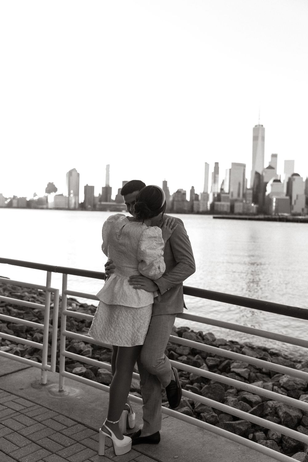 Silhouetted couple admires Manhattan skyline while embracing on riverside walkway.
