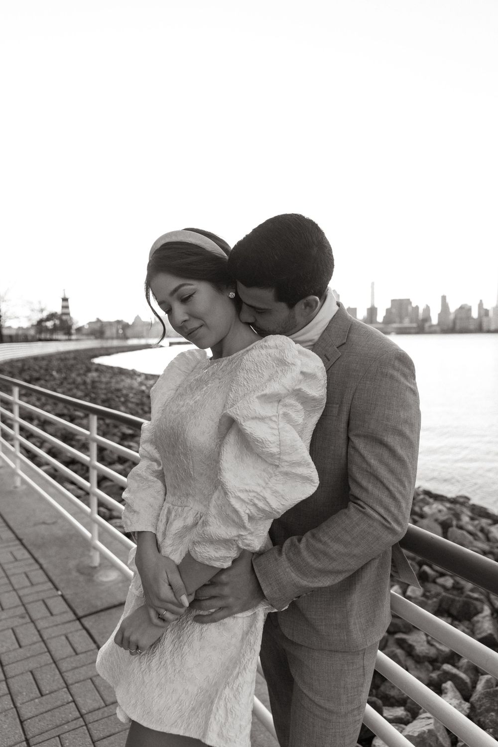 Intimate black and white portrait of couple embracing by waterfront with city backdrop.