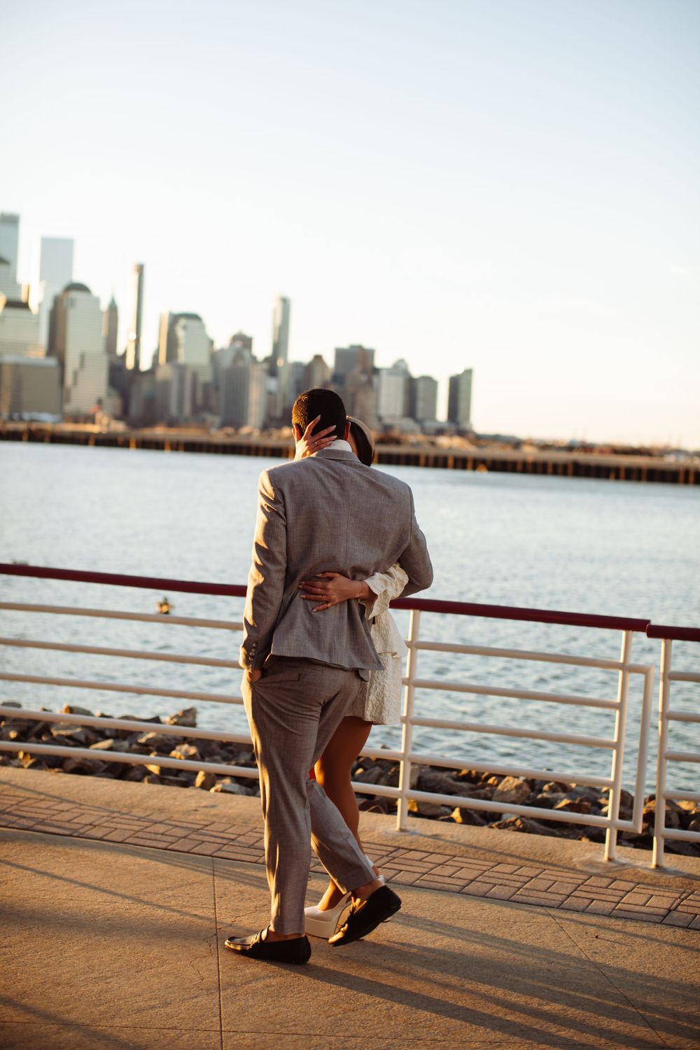 A couple embraces while walking along a waterfront promenade with the New York City skyline in the background at sunset.