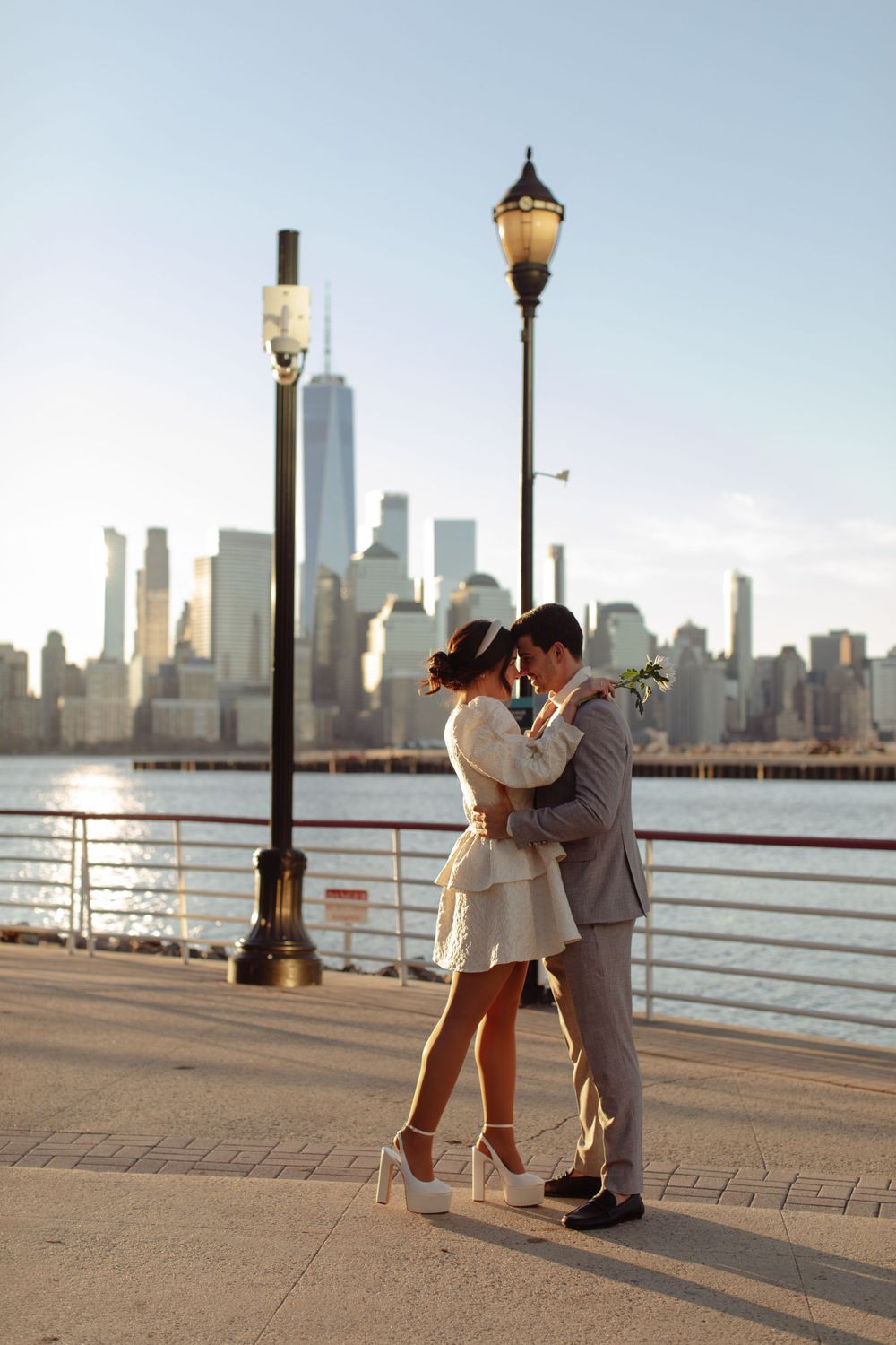 Romantic couple embracing by vintage lamp posts along river with Manhattan skyline view.