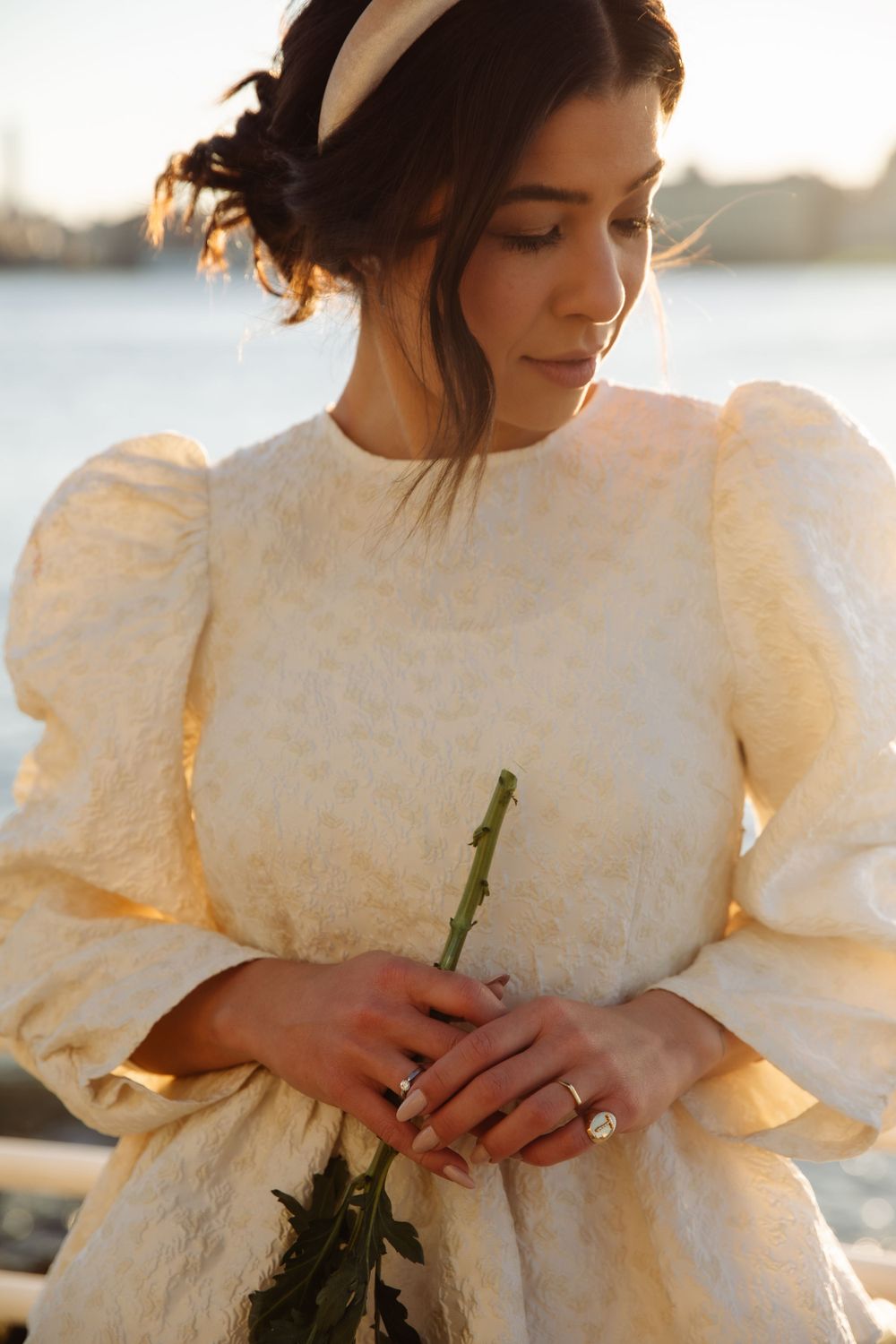 Bride in vintage-style white dress with puff sleeves holding flower against sunset waterfront.