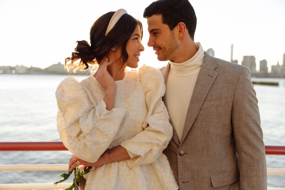 Romantic couple in wedding attire embrace by waterfront with city skyline visible.