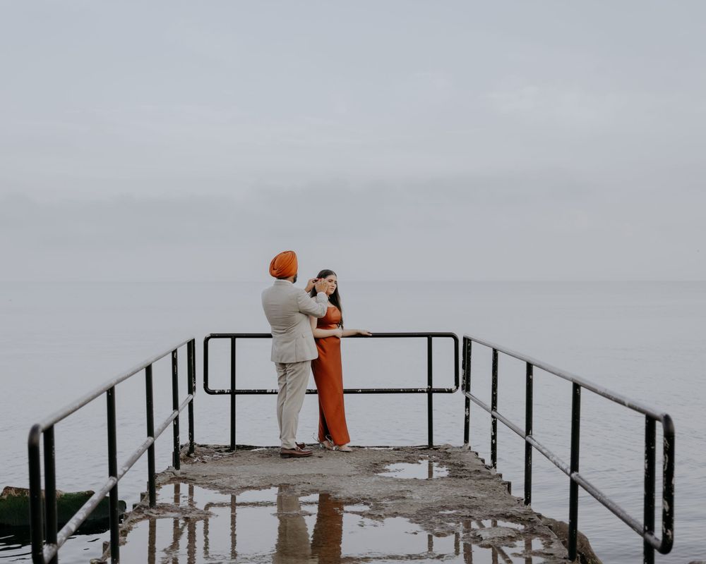 A couple embraces on a wet pier with metal railings against a cloudy sky.
