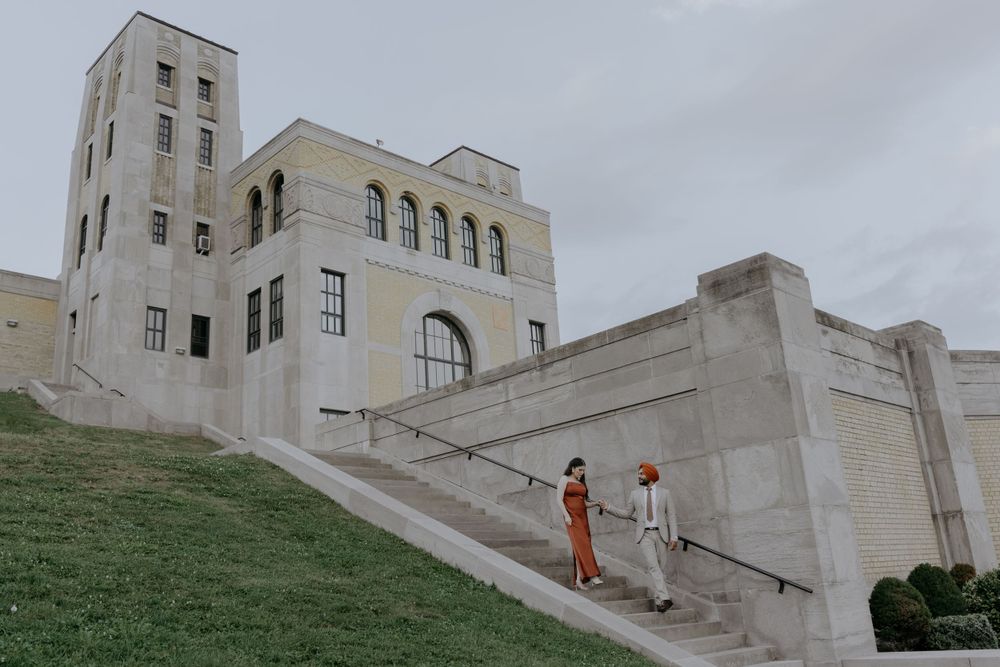 A couple ascends white stone steps of an architectural landmark with arches and towers.