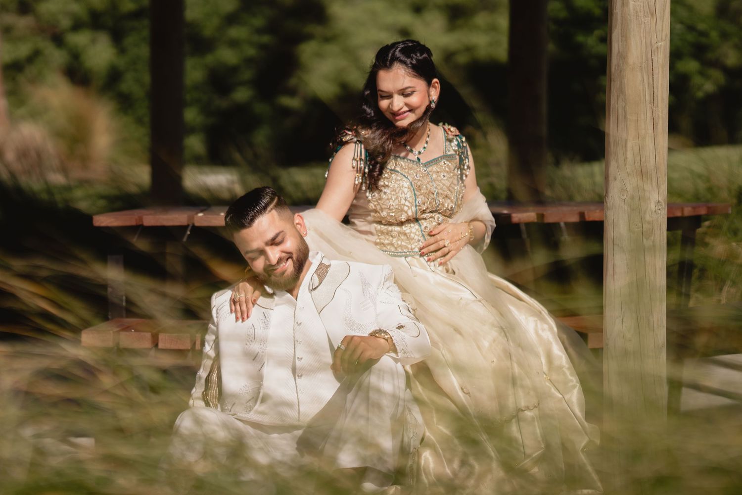Joyful South Asian couple in traditional wedding attire sharing a playful moment outdoors among lush greenery.