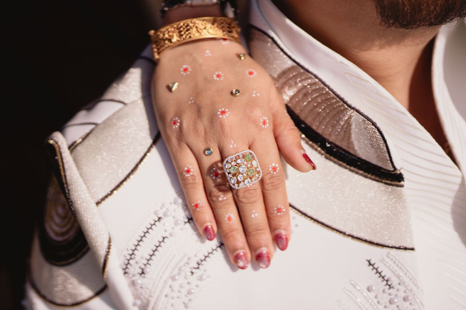 Close-up of a hand wearing a large diamond ring and gold bracelet, with decorative henna dots and red nail polish.