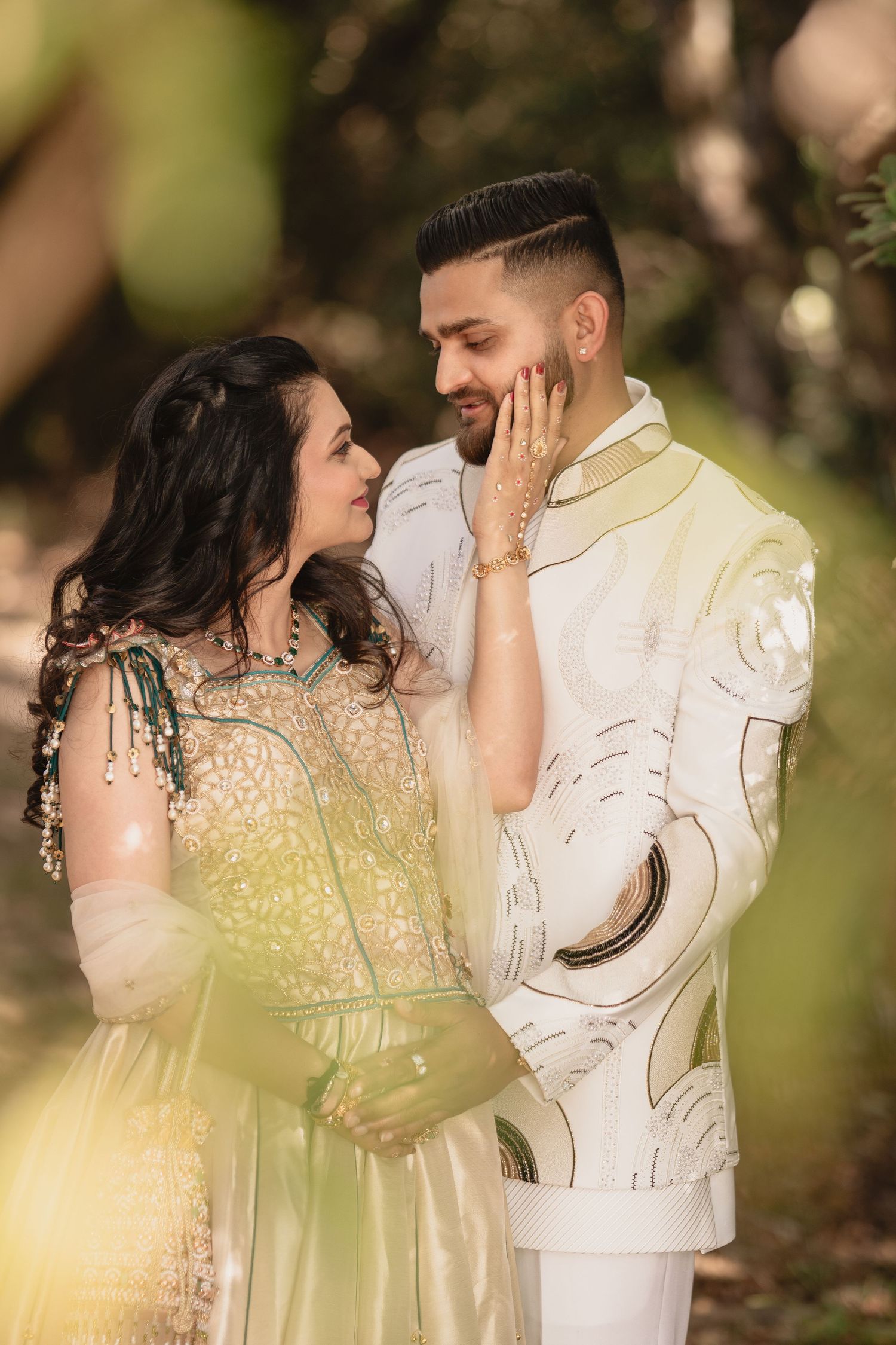Couple in elegant Indian wedding attire, woman in green lehenga touching mans face, sharing romantic moment outdoors.