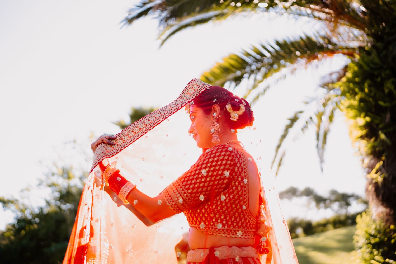 Bride in red embroidered lehenga and dupatta posing outdoors near palm trees on her wedding day.