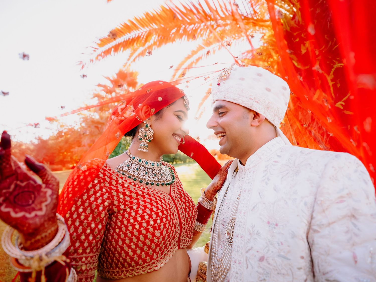 Indian bride in red lehenga and groom in white sherwani sharing a joyful moment at traditional Hindu wedding.