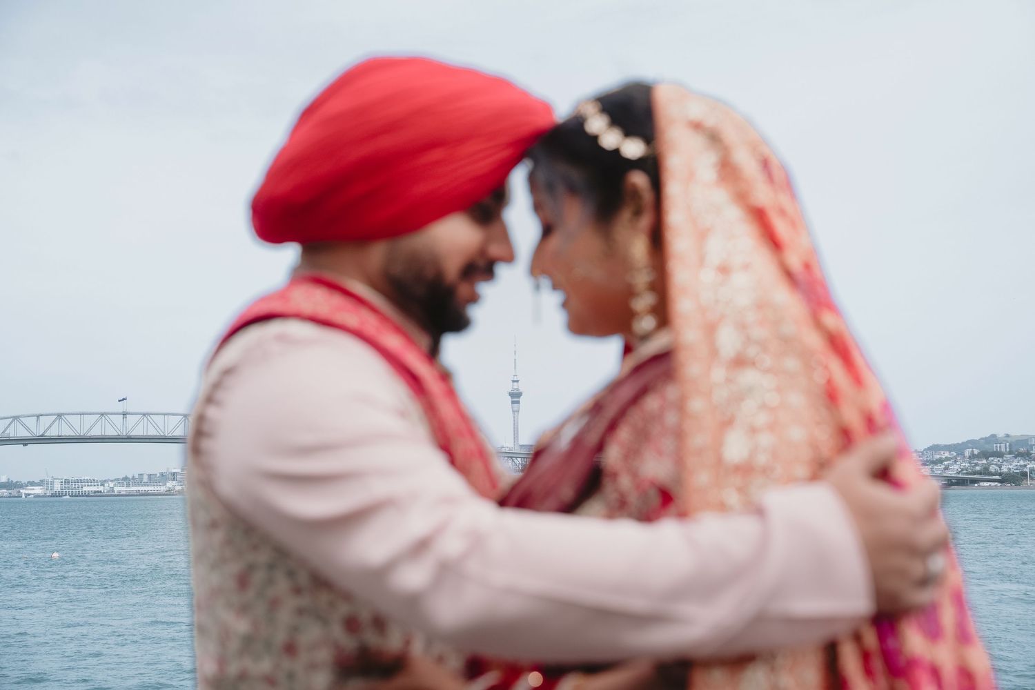 Sikh couple in traditional wedding attire embracing near waterfront with Auckland Sky Tower in background.