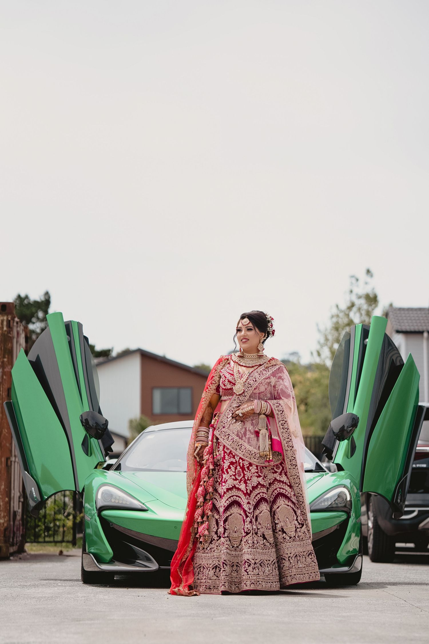 Indian bride in red lehenga poses before a green McLaren supercar with scissor doors open on her wedding day.