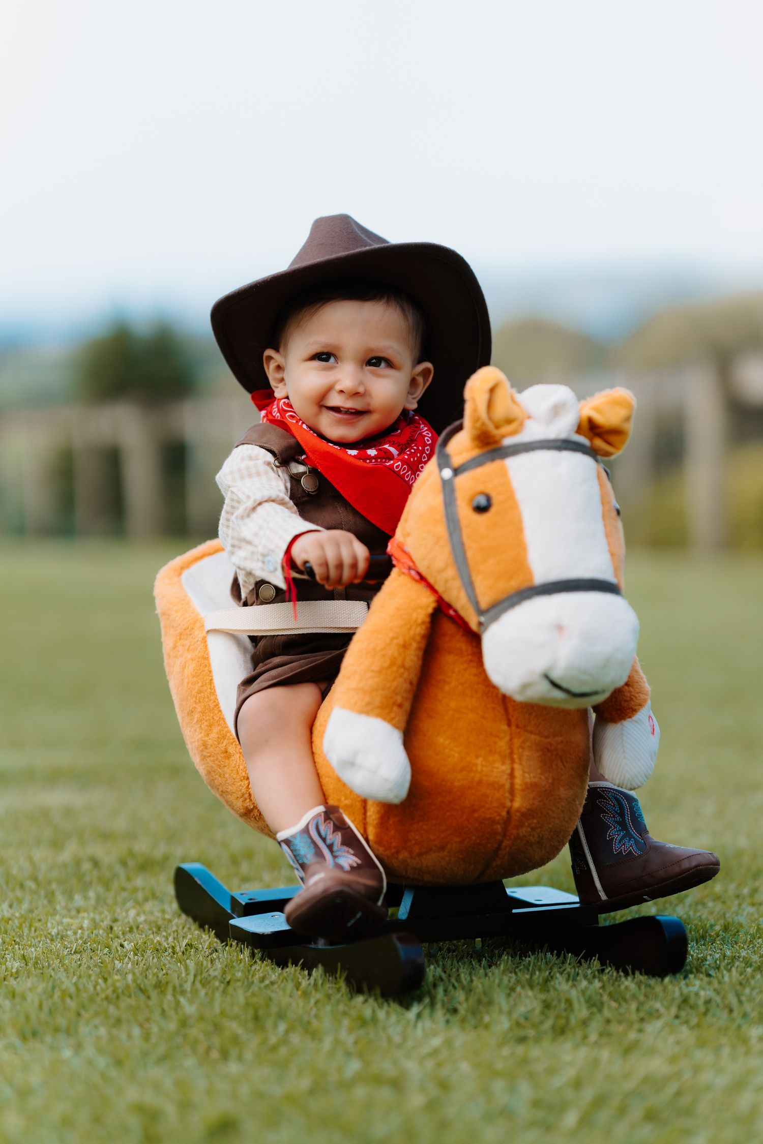 Adorable baby dressed as a cowboy with hat and bandana, sitting on an orange plush rocking horse on green grass.
