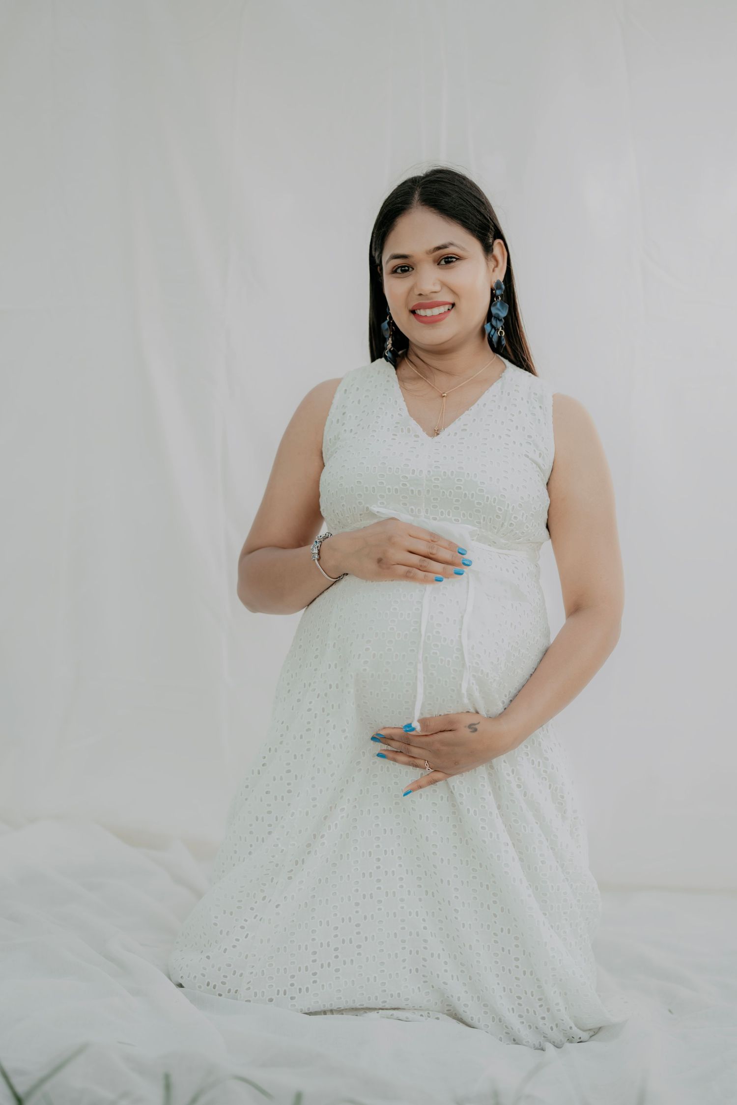 Maternity portrait featuring woman in white eyelet dress against neutral backdrop.