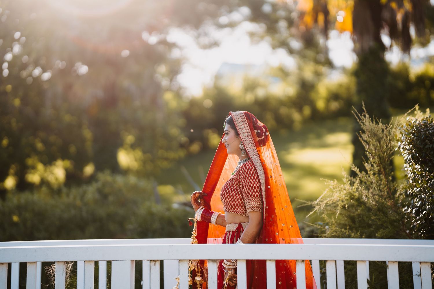 Indian bride in vibrant orange and red lehenga with dupatta, standing on white bridge surrounded by lush greenery.