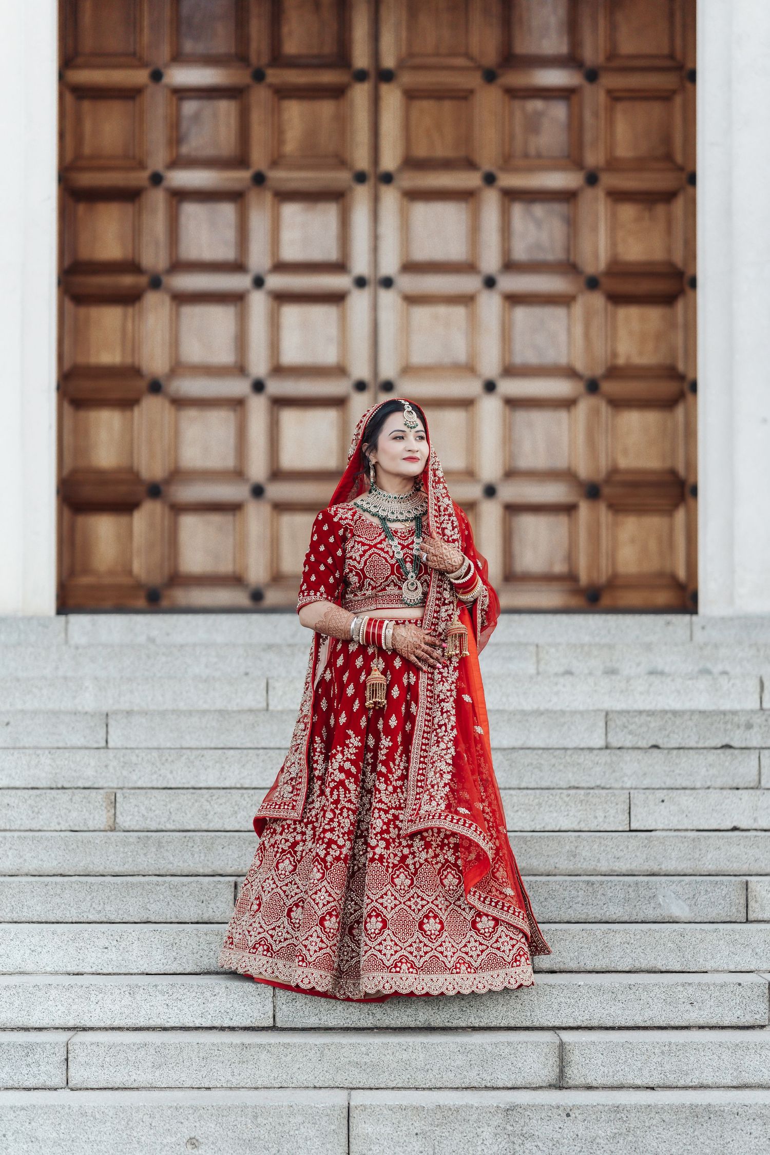 Beautiful traditional red bridal lehenga with intricate embroidery photographed on stone steps with wooden door backdrop.