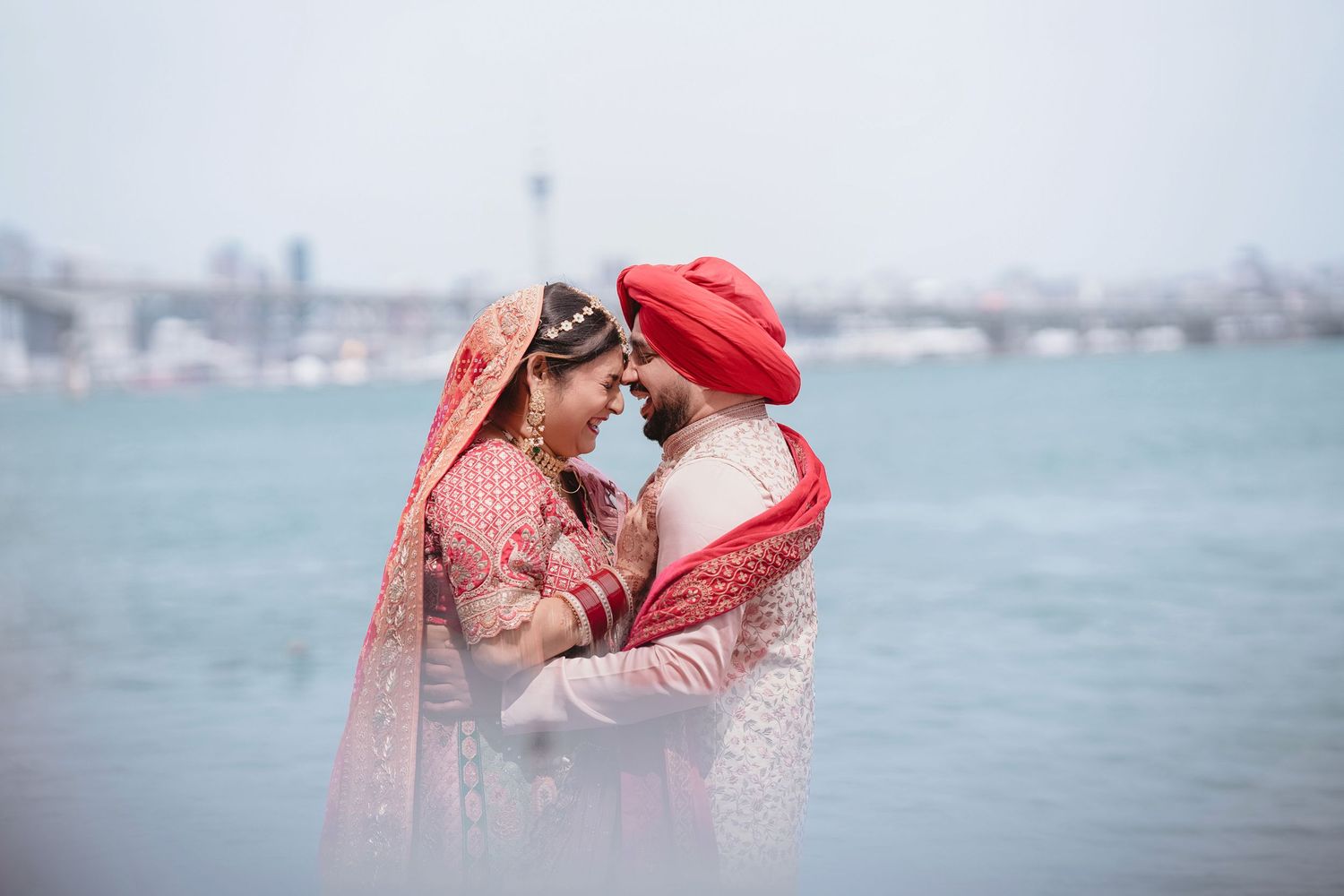 Indian couple in traditional red wedding attire embracing by the waterfront, bride in lehenga and groom in turban.