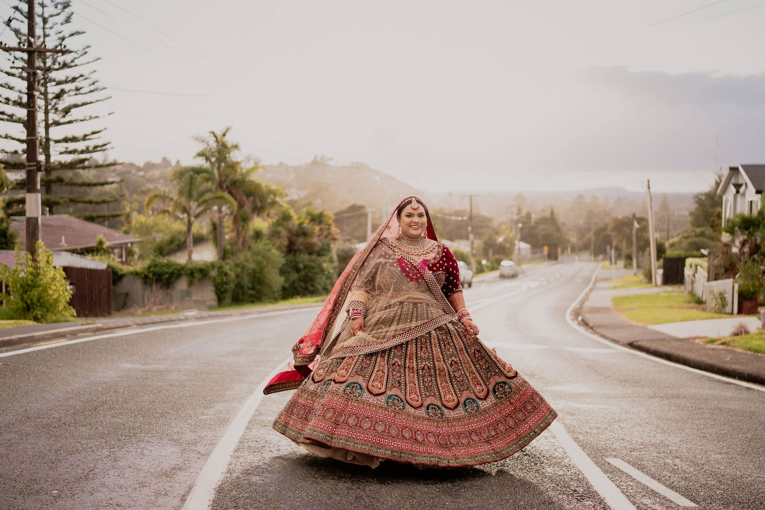 Bride in vibrant red and gold lehenga twirling on a wet road with lush green trees and hills in background.