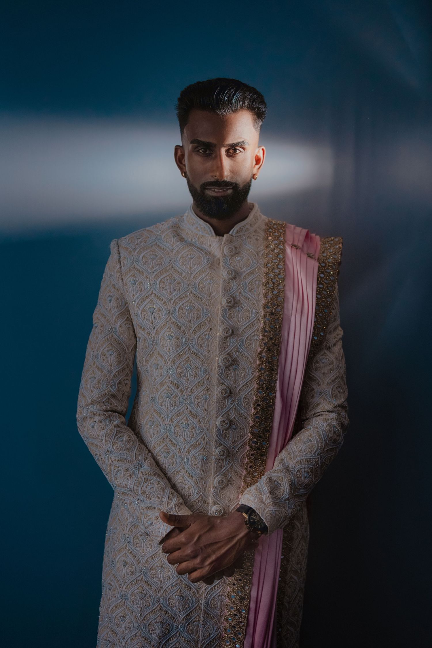 Man in embroidered cream sherwani with pink dupatta posing against teal background for Indian wedding.