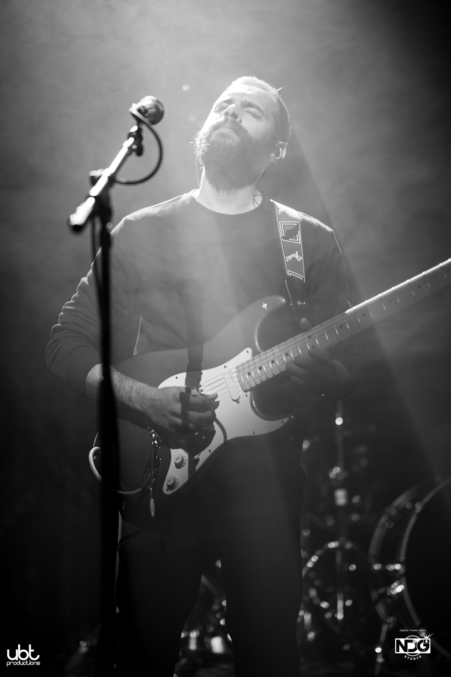 Dramatic black and white concert photo of a musician playing electric guitar under spotlights.