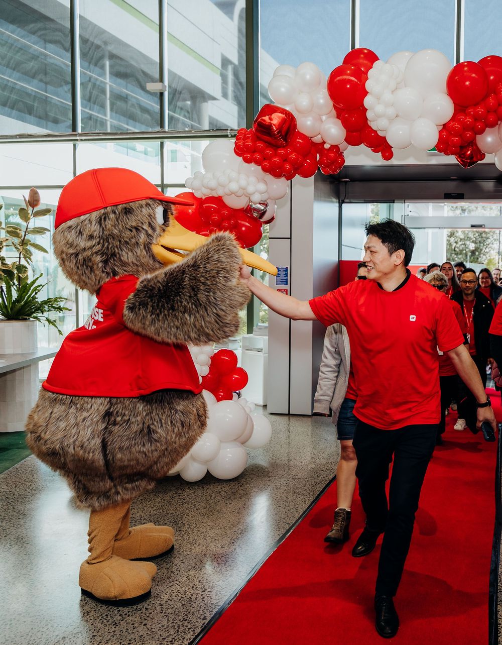 A man in red uniform high-fives a large bird mascot on a red carpet surrounded by red and white balloons.