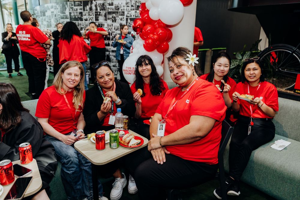 Group of women in red shirts smiling at a corporate event with balloons and drinks on a tray.