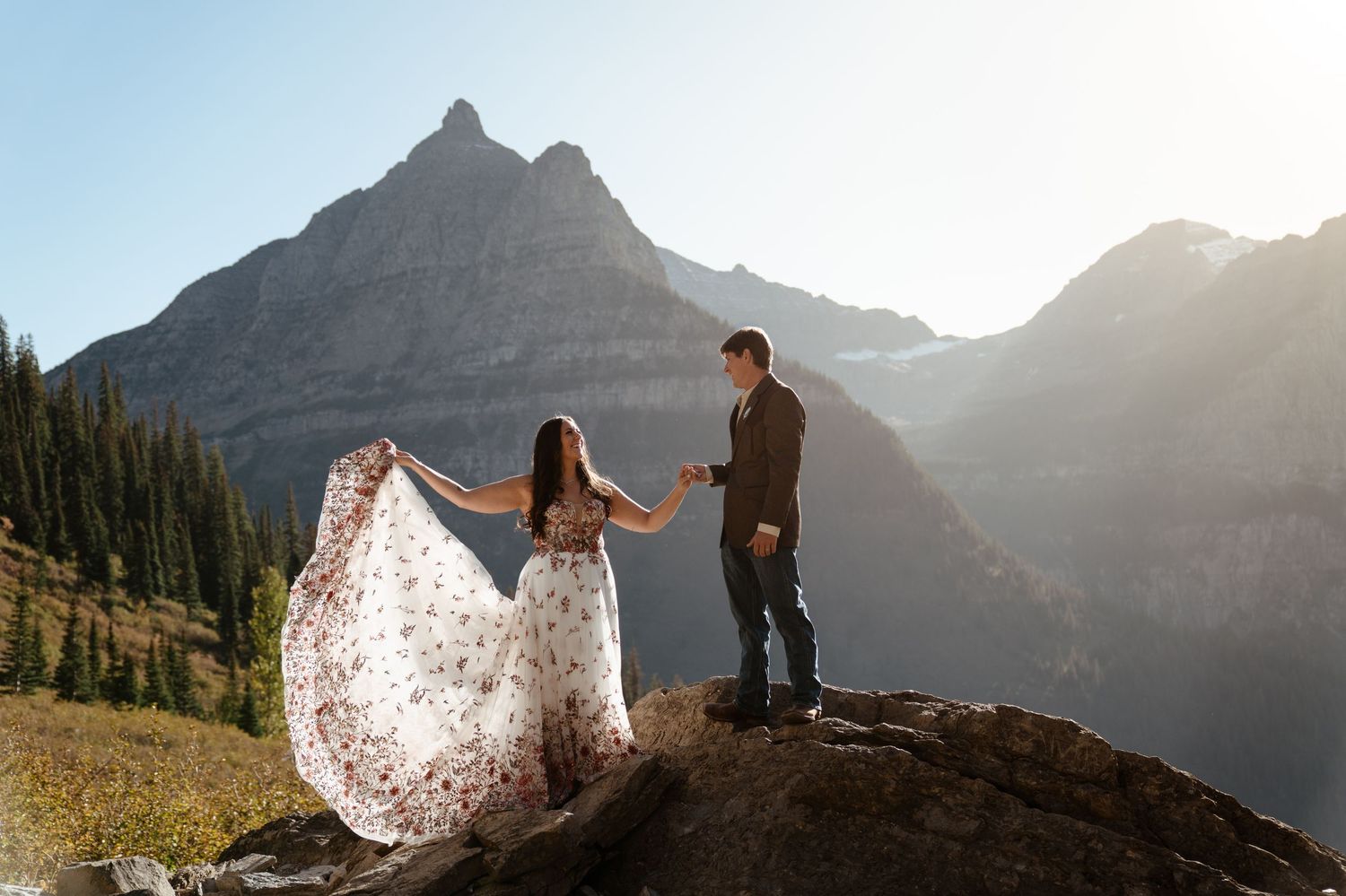 Bride wearing colorful wedding dress with red embroidered florals sways dress in the wind while holding grooms hand in Glacier National Park