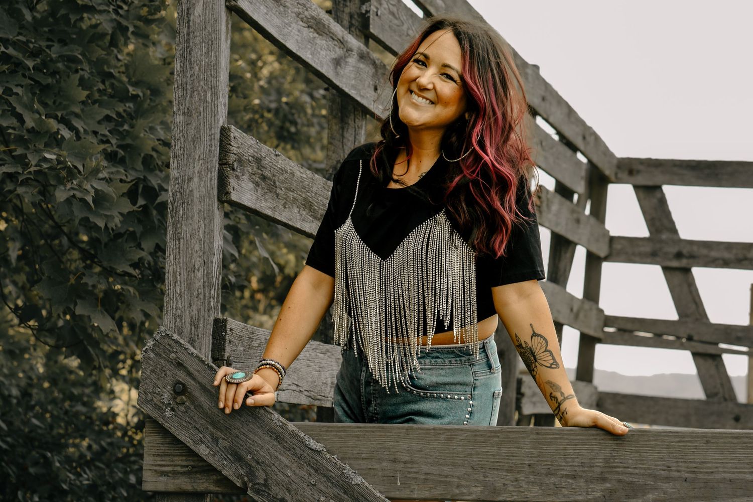 Woman wearing black fringed top leans on wooden railing against autumn foliage background.