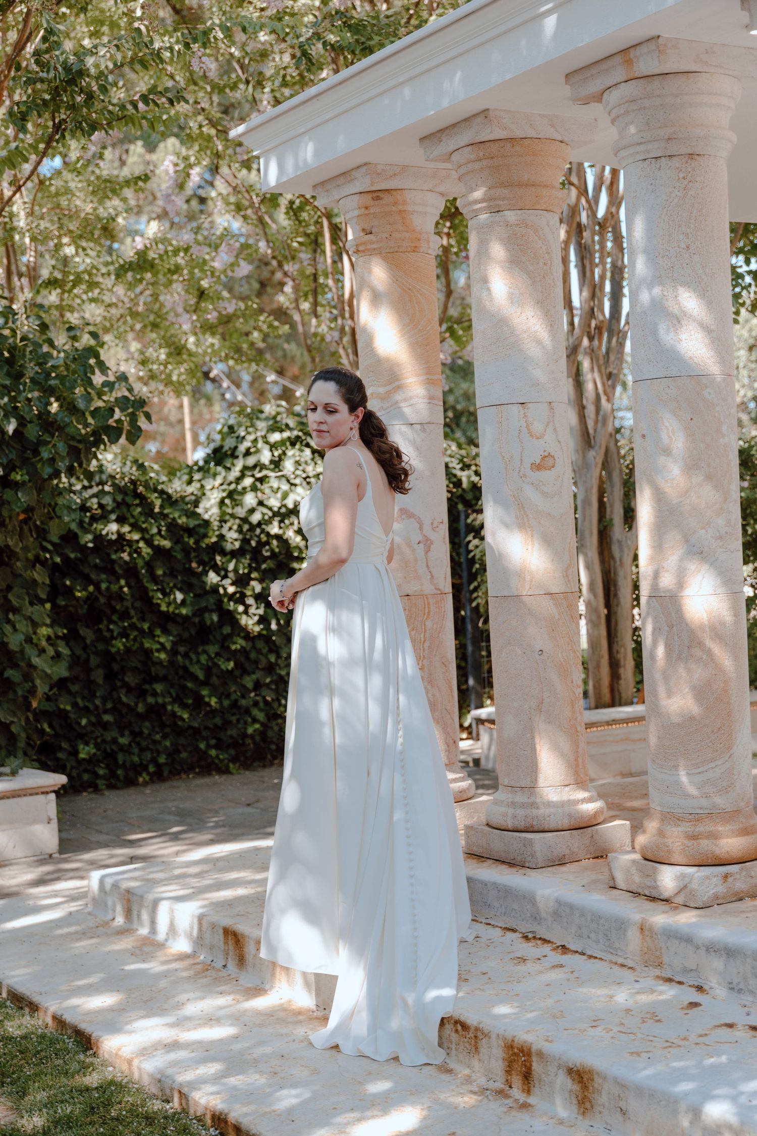 Elegant bride in flowing white dress poses by classical stone columns in garden setting.