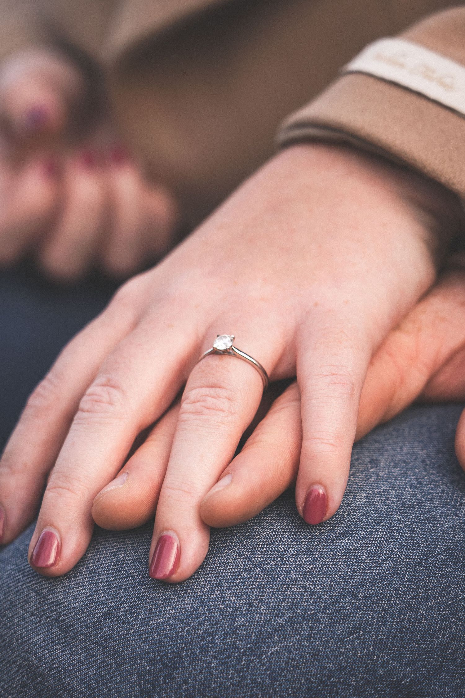Close-up view of hands with burgundy nail polish and a wedding ring resting on dark gray fabric.