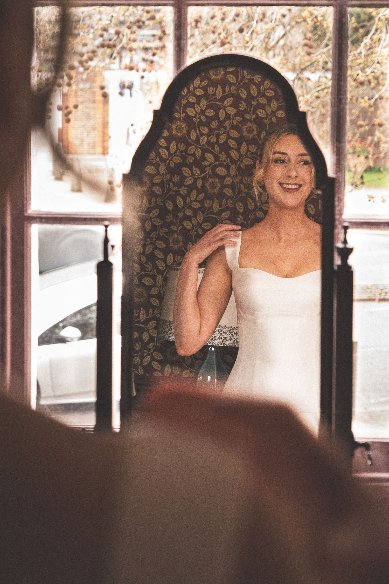 Smiling bride in white wedding dress standing in ornate doorway with floral arch detail.