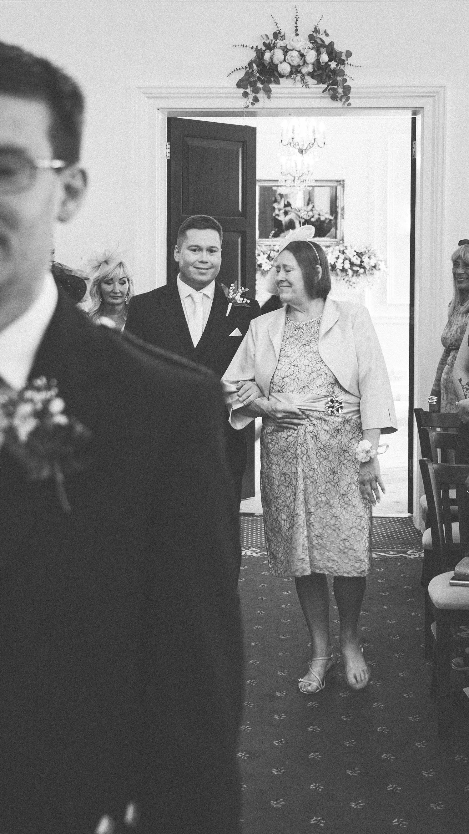 Black and white photo of a man in a suit escorting an older woman down the aisle at a wedding ceremony.