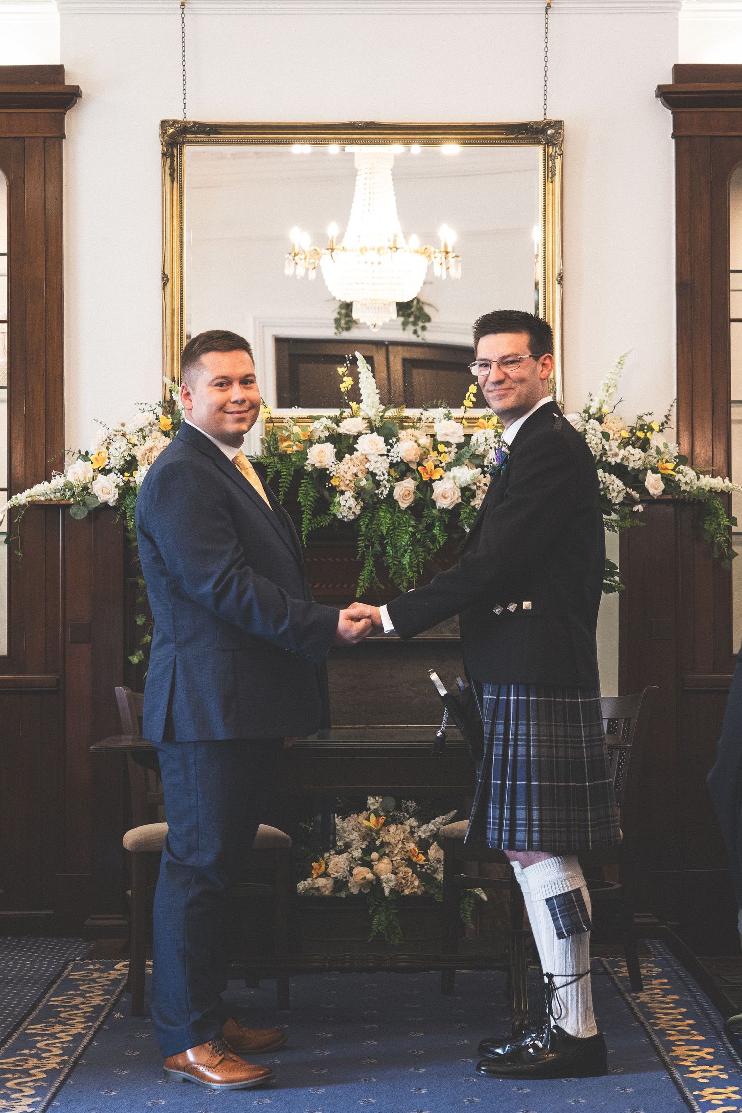 Two grooms shaking hands at wedding ceremony, one in navy suit, one in traditional Scottish kilt, with floral arrangements.