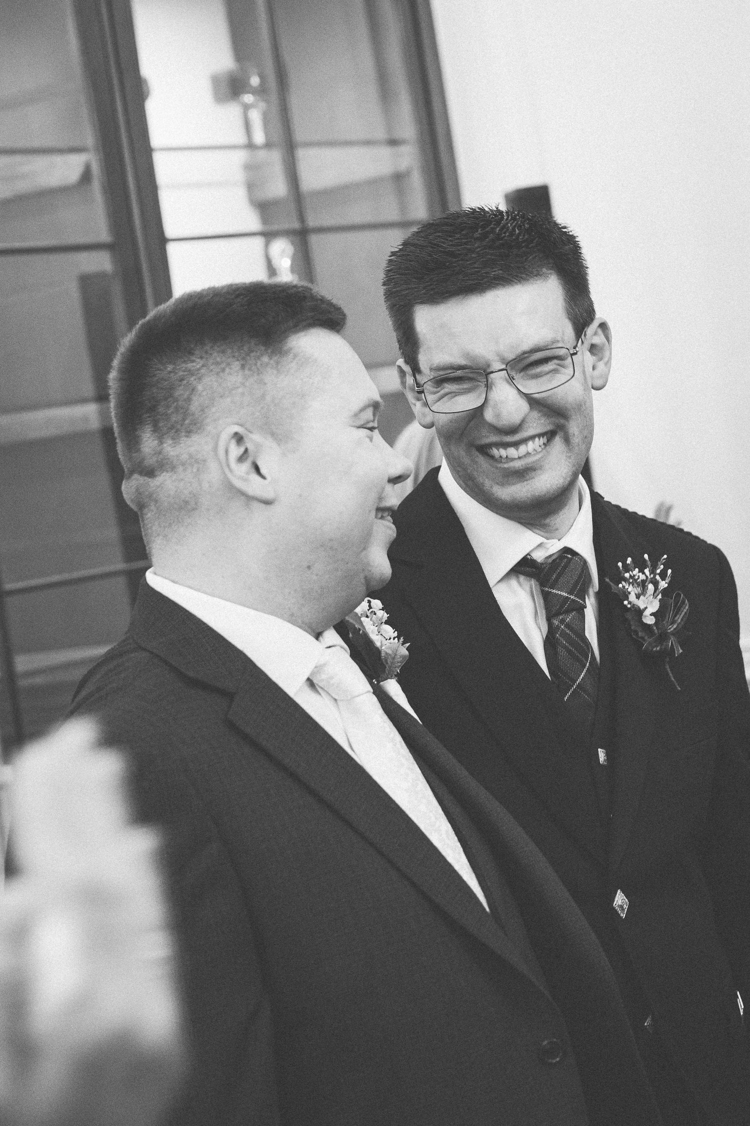 Two grooms share a joyful moment at their wedding, smiling at each other in suits with boutonnieres.