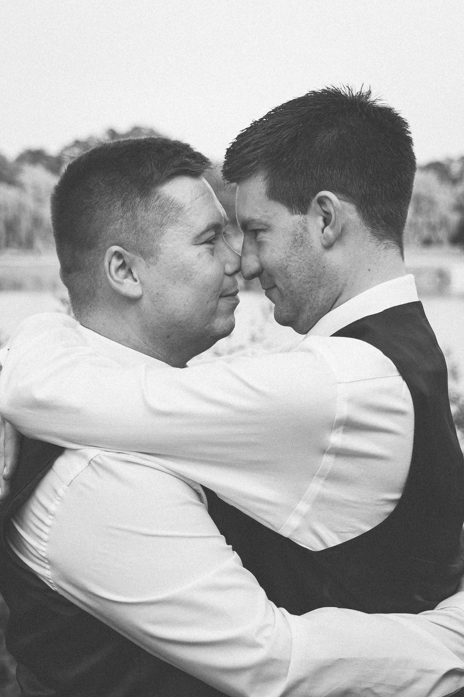 Black and white photo of two grooms in vests embracing closely, foreheads touching, at their wedding.
