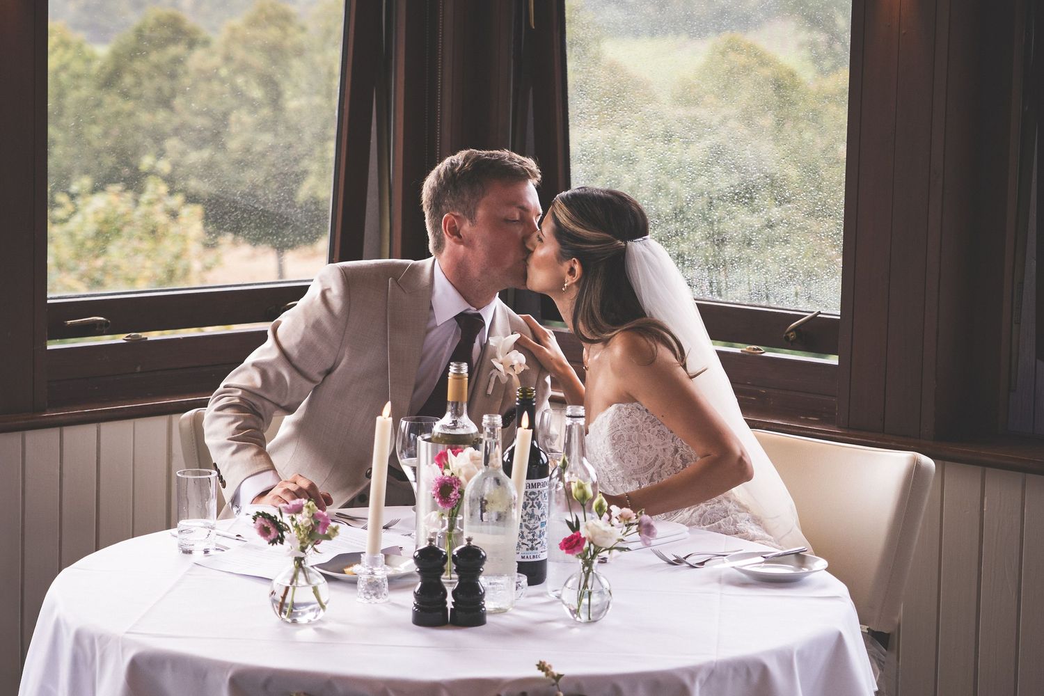 Bride and groom share a kiss at elegant wedding reception table with candles and champagne glasses.
