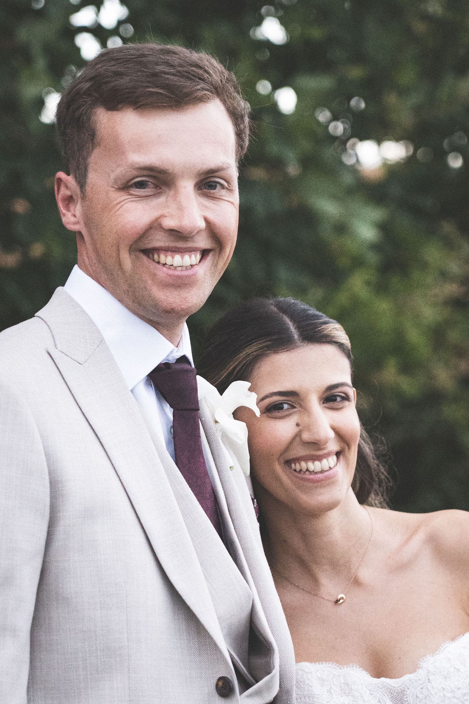 Smiling bride and groom in light gray suit with burgundy tie posing outdoors on their wedding day.