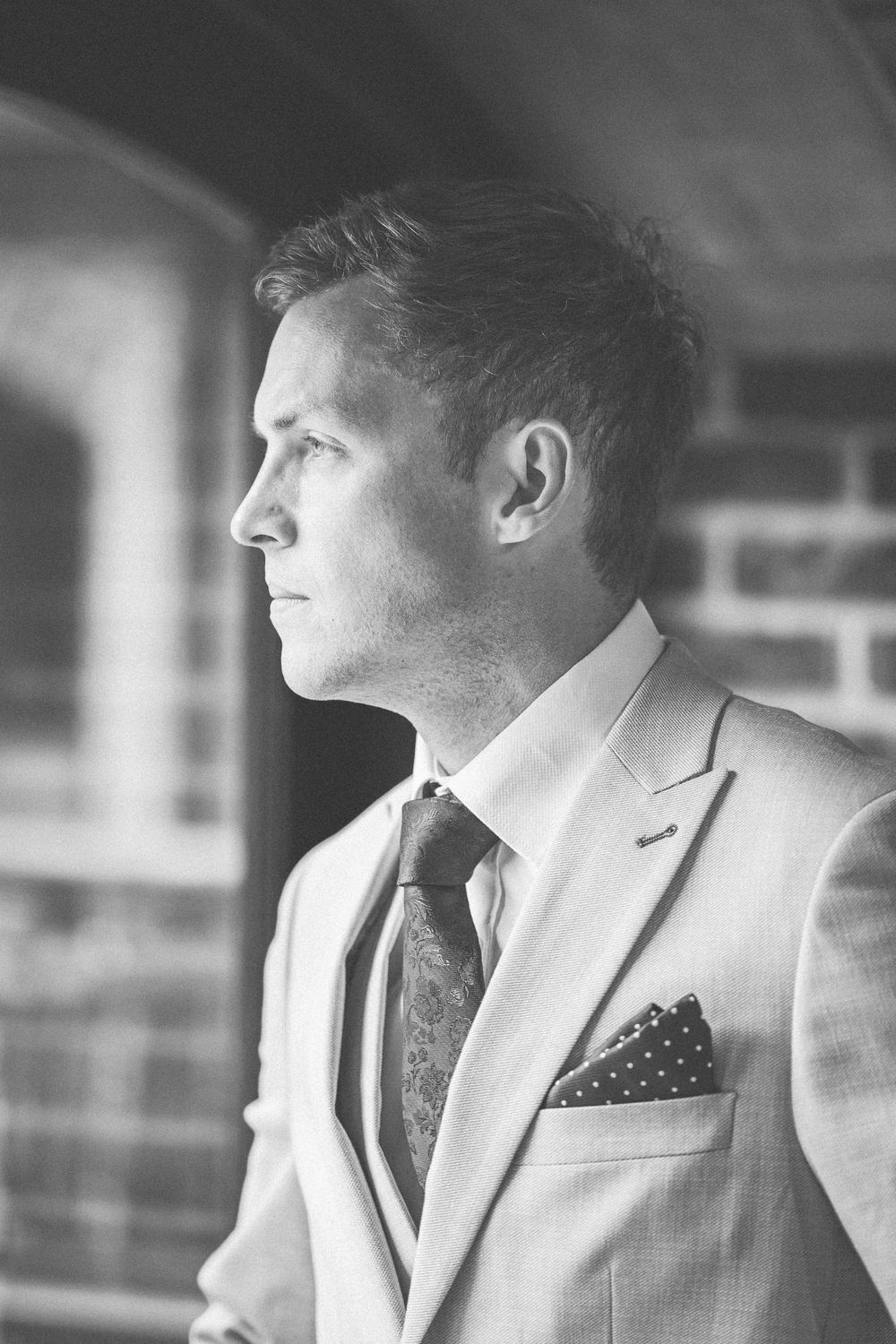 Black and white portrait of a well-dressed man in a suit and tie with pocket square, gazing thoughtfully to the side.