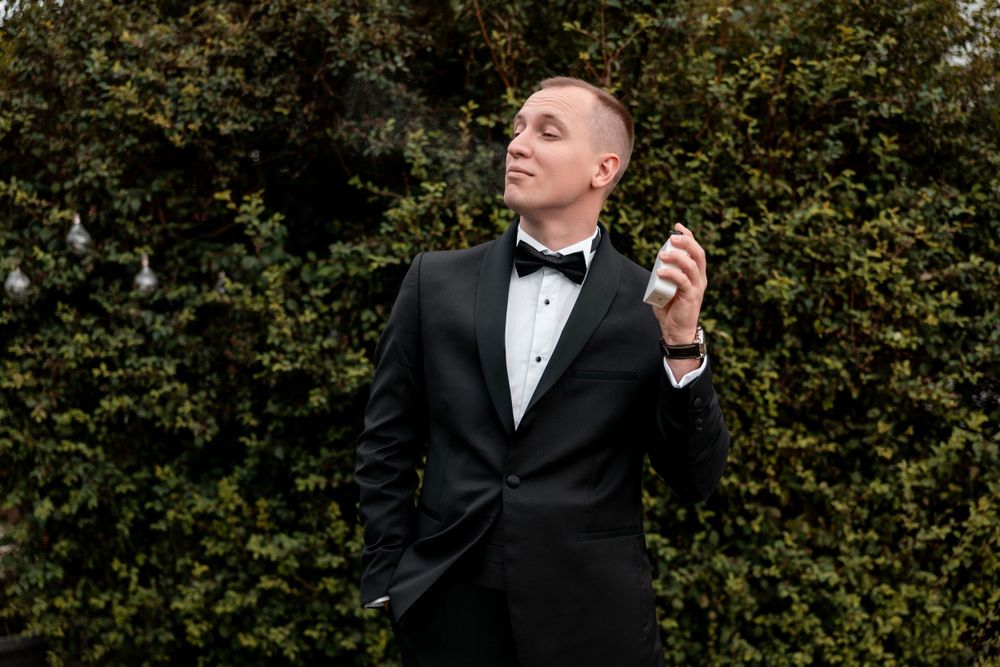 A groom in classic black tuxedo poses thoughtfully against garden foliage.