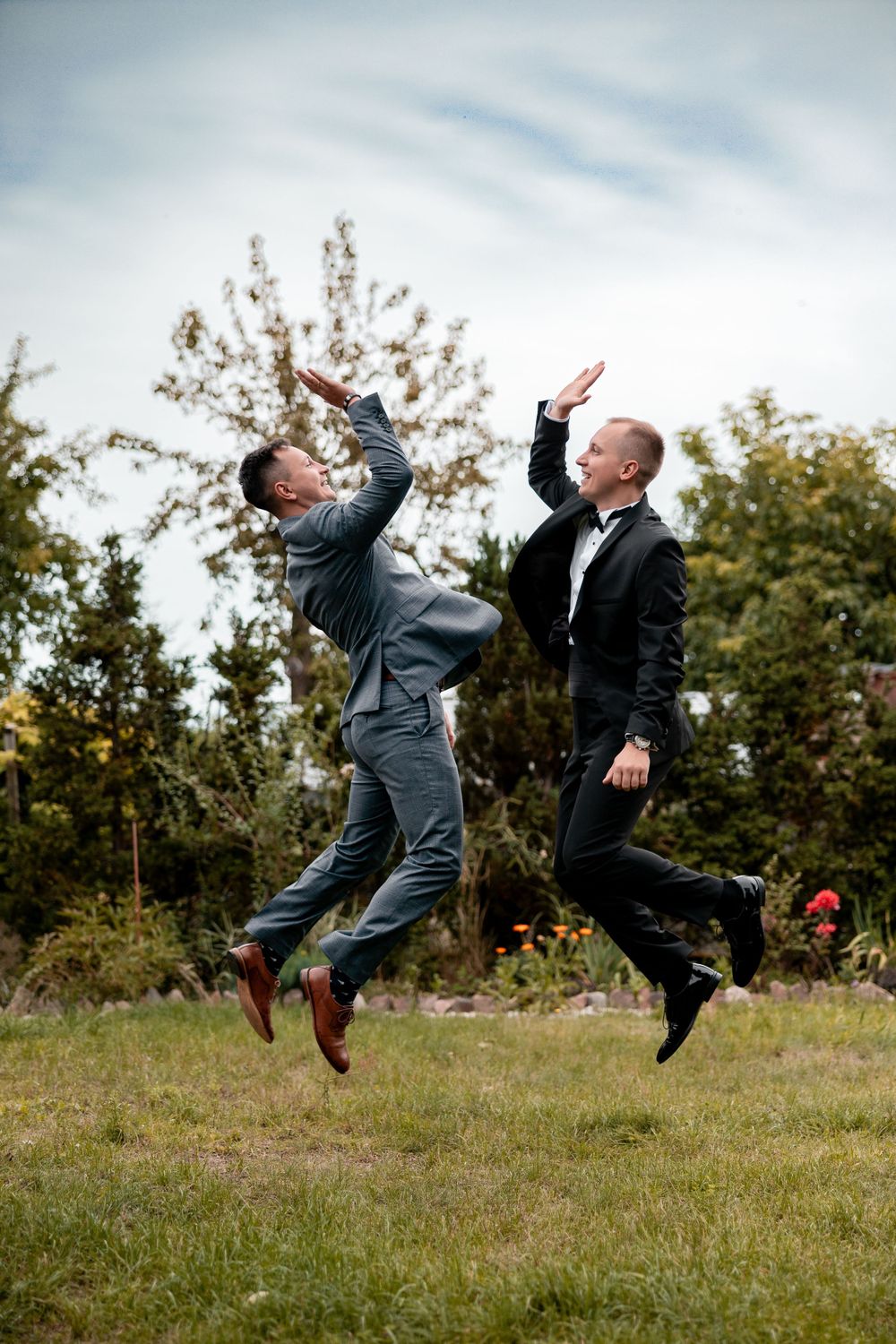Two elegantly dressed men jumping playfully on grass during a wedding celebration.