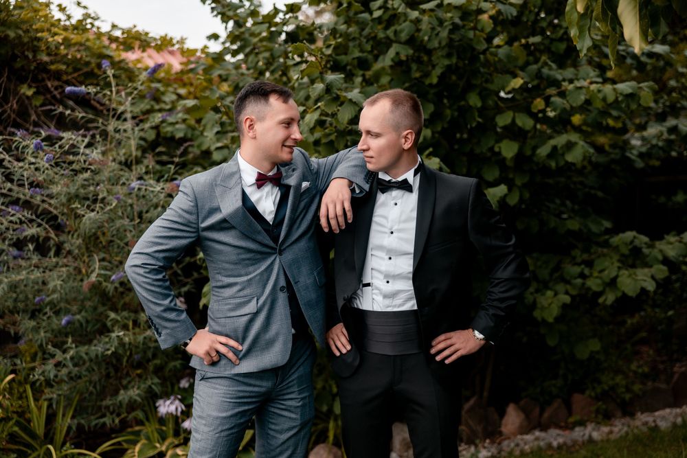 Two wedding guests share a friendly moment in a garden setting.