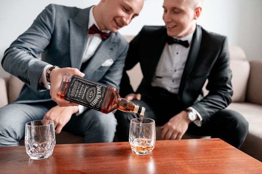 Two people in formal suits pouring whiskey into crystal glasses on a wooden table.