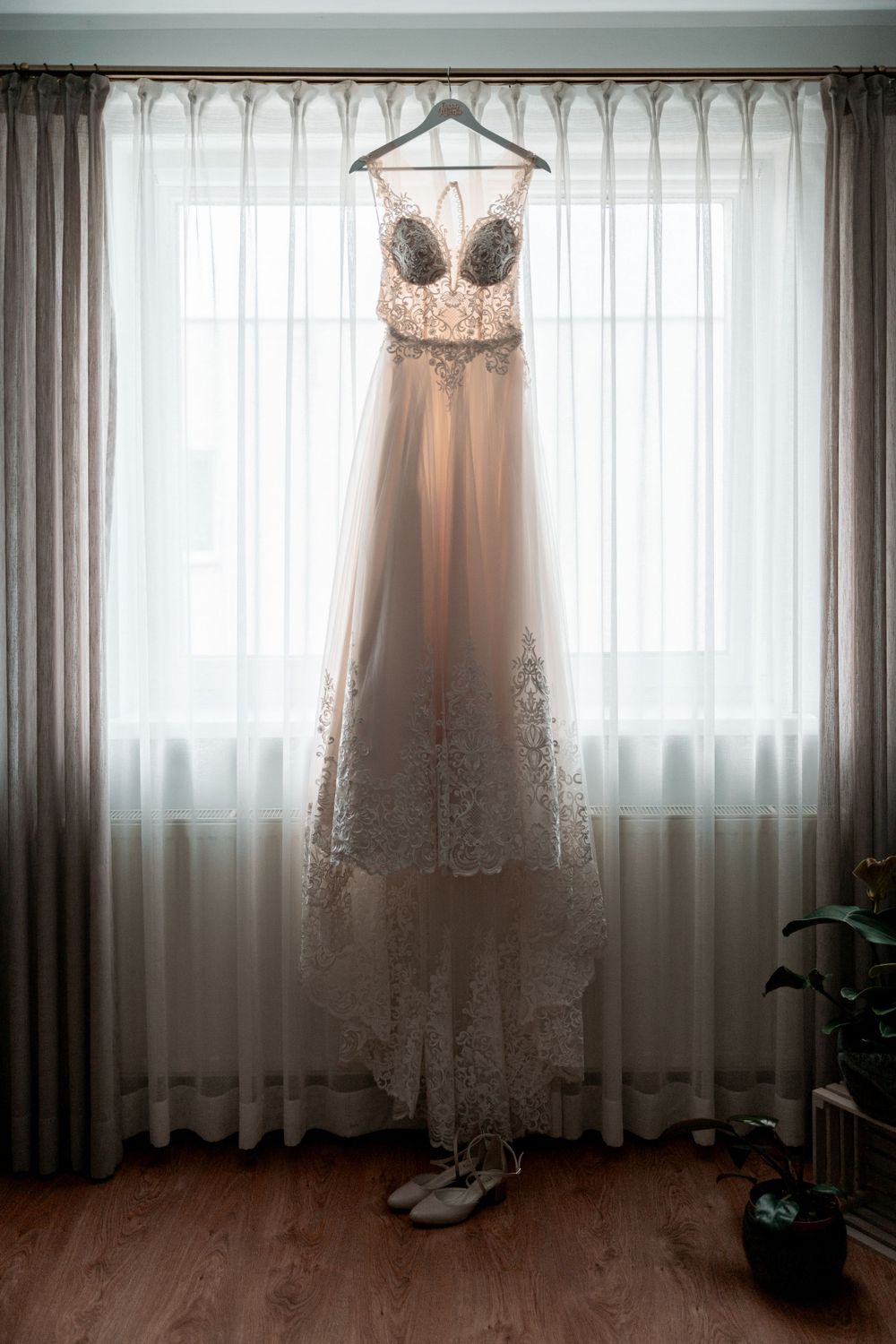 Elegant lace wedding dress hanging on display against sheer curtained windows.