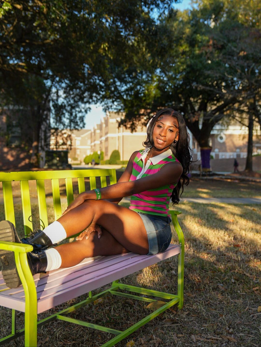 Woman in a striped green and pink top, denim skirt and black boots sits on a bright green bench in a sunny campus park.