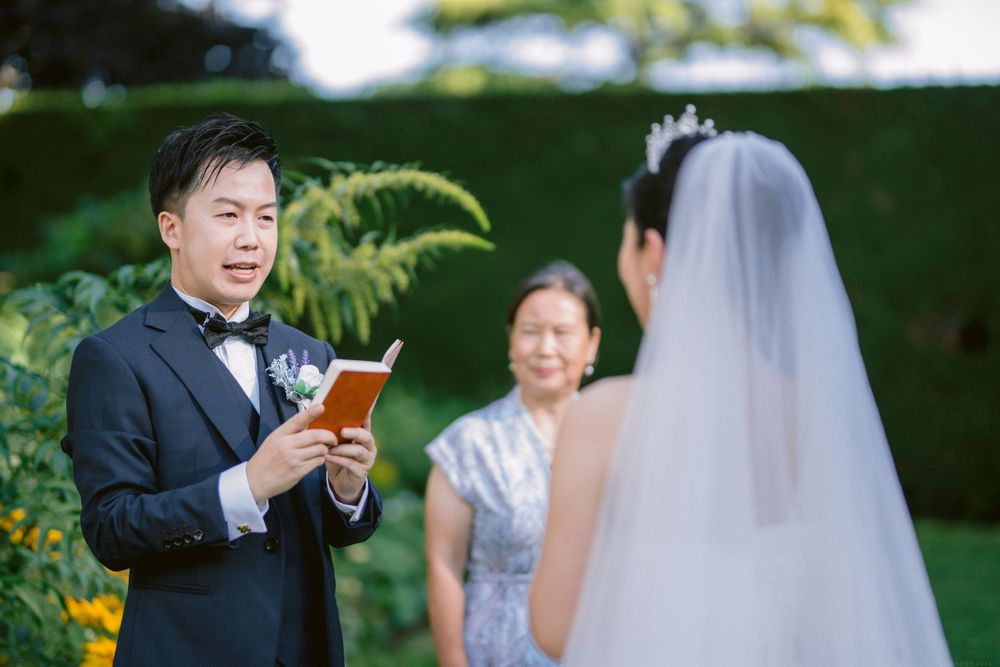 A wedding ceremony in a garden where the groom reads vows from a card while wearing a black tuxedo.