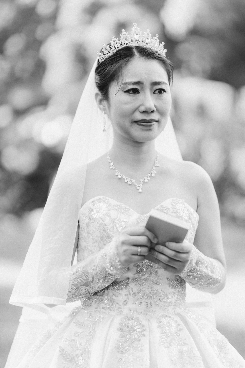 Elegant bride in a white lace ballgown and tiara holding a card during an outdoor wedding ceremony.